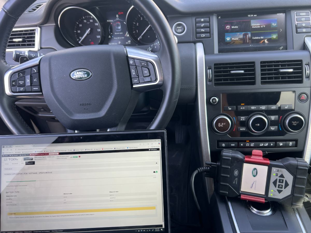 Land Rover Discovery Sport interior with Land Rover steering wheel and a laptop running JLR TOPIx Diagnostics for a PCM software update, branded Autel scan tool plugged in via OBD-II